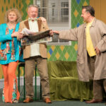 na pierwszym planie kobieta trzyma mężczyzna za ramię, mężczyzna czyta z uśmiechem gazetę. po prawej stoi drugi mężczyzna w płaszczu który łapie pierwszego za rękę, w tle okno z widokiem na drugi budynek