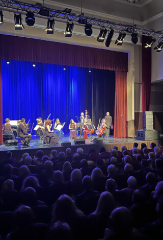 na pierwszym planie widownia, na drugim od lewej fortepianistka, sześć skrzypków i dwóch wiolonczelistów